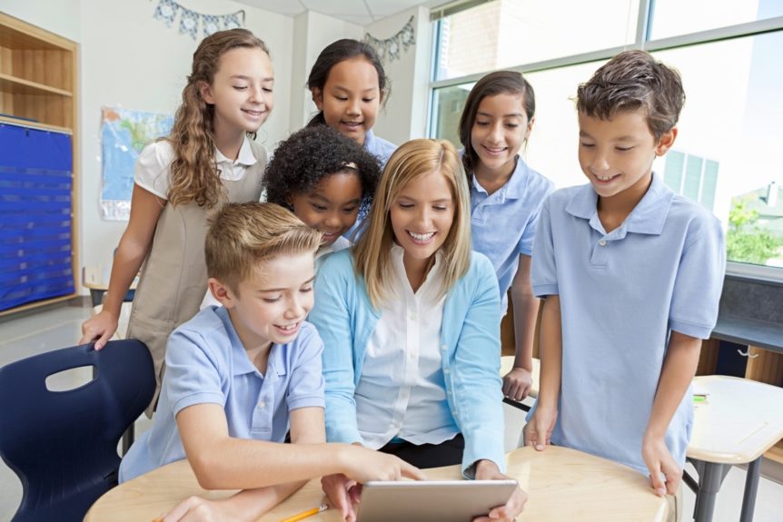 Caucasian mid adult female teacher uses a digital tablet to show a new class project to her multi ethnic students. The group is sitting and standing with their teacher at a desk in the classroom. The students are happy and smiling.